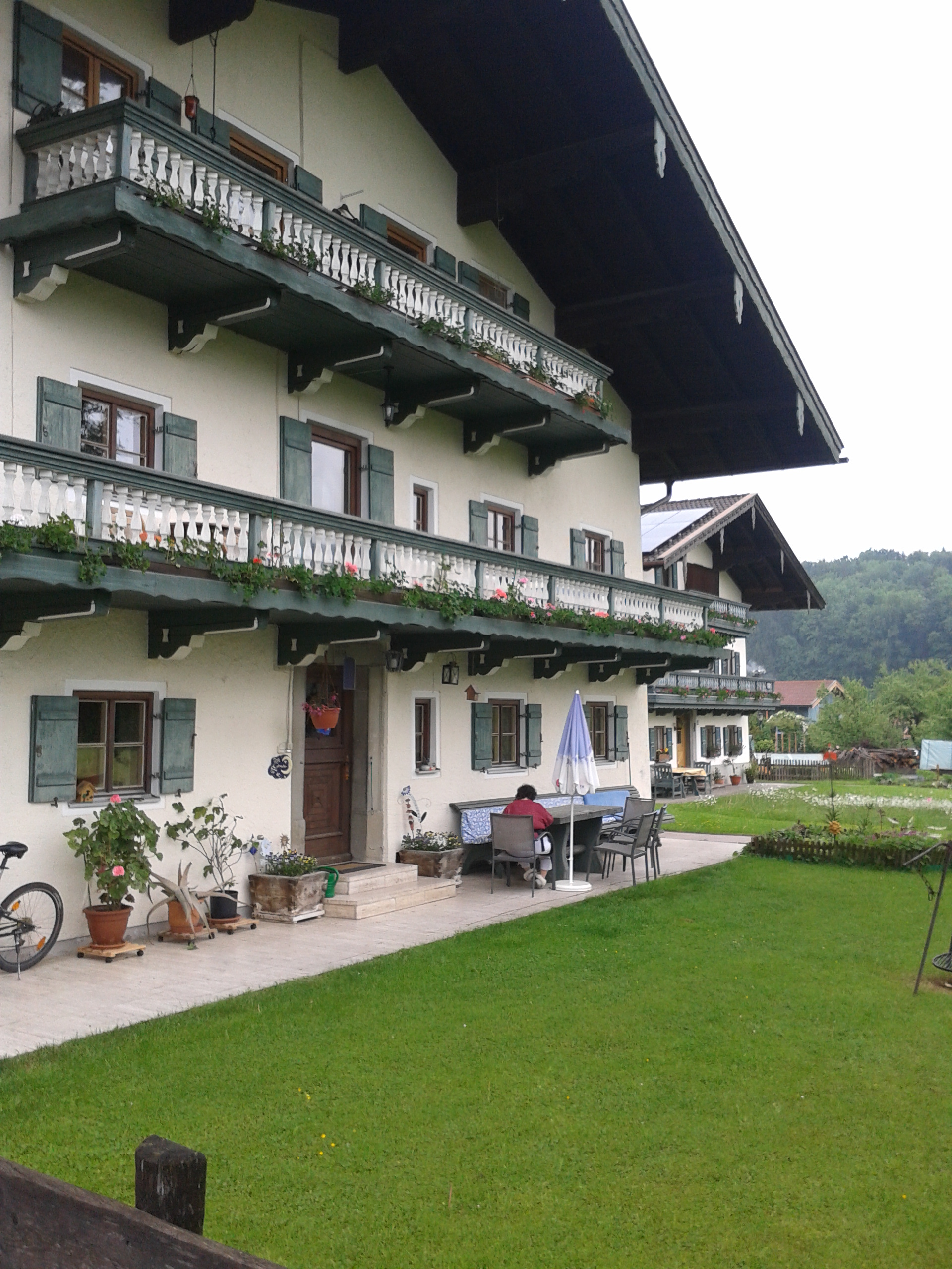 Das holzverkleidete Bauernhaus in seitlicher Aufnahme,links Blumentöpfe,rechts Tische,Stühle und Bänke. Darüber mehrere Balkone und kleine Fenster. Der ganze Bauernhausbereich ist überdacht und von einem Holzzaun begrenzt. Vor der Terrasse eine Wiese mit Grillplatz.